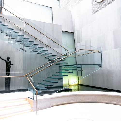 Side view of the all-glass staircase at the Onassis Cultural Center showcasing the stairs, ballustrades and railing.